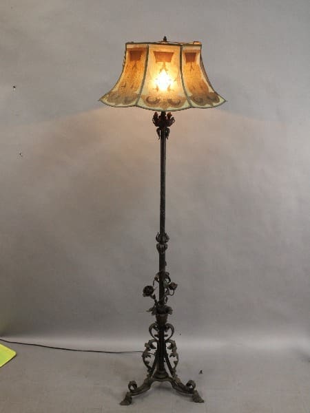 An antique 15297. Spanish Revival Wrought Iron floor lamp with a detailed metal base and a lit, bell-shaped shade, set against a plain backdrop.