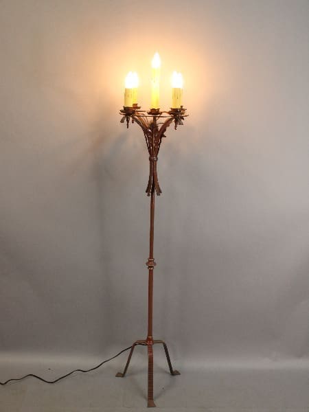 A tall 15639. Spanish Revival floor lamp with a wooden stand and three electric candles, casting a warm light against a plain, light background.