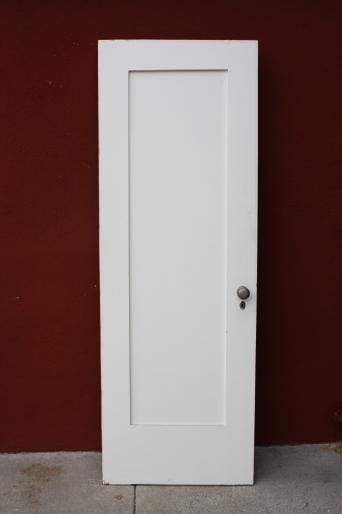 A white, 6554. 1920s Single Panel Interior Door GG with a round doorknob, standing against a red wall on a grey concrete floor.