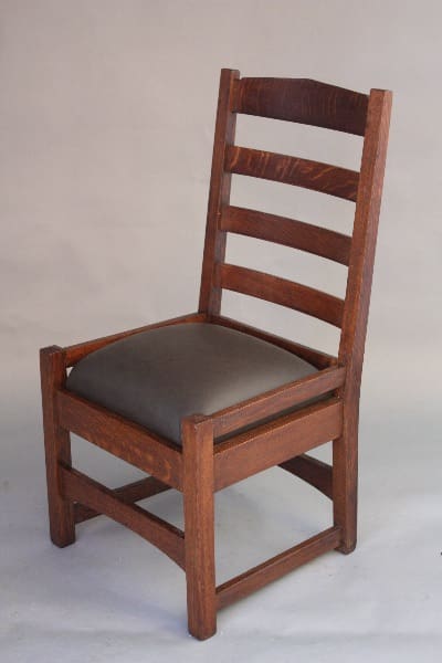 8455. Arts And Crafts Oak Ladder Back Chair with a dark brown finish and a padded, black leather seat cushion, against a neutral gray background.
