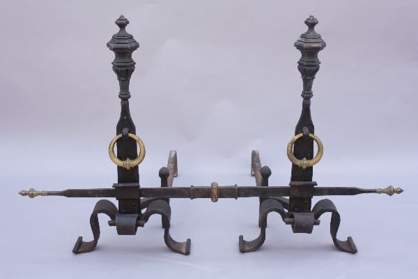 Antique bronze and wrought iron fireplace andirons with intricate designs and ring tops on a plain background.