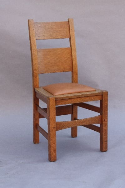 A simple wooden 9780. Arts & Crafts Period Side Chair with a tan cushioned seat against a grey background.