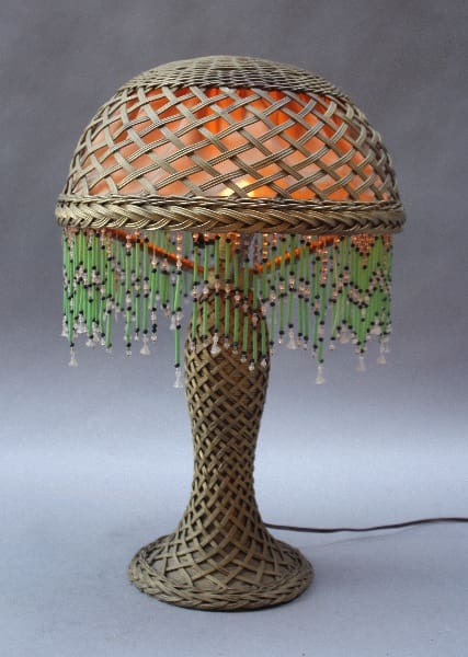 A 9875. Arts & Crafts Wicker Table Lamp w/ Glass Bead Fringe with a dome-shaped top and bead fringes, illuminated from within, standing against a gray background.