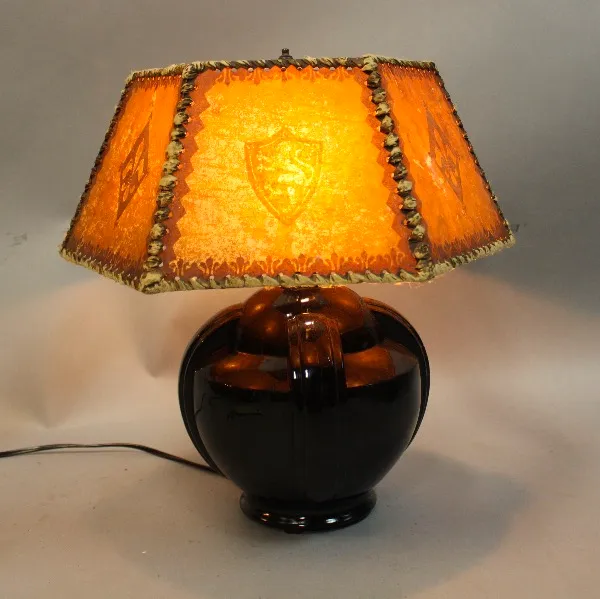 A 15913. Antique Mica Lamp with Pottery Base with an ornate orange shade featuring a shield design, resting on a glossy black spherical base, illuminated.
